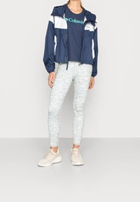 Granatowo-biała kurtka wiatrówka na zamek z kapturem, zestawiona z granatowym t-shirtem z zielonym logo oraz jasnymi legginsami w kwiatowy wzór. Beżowe tenisówki.