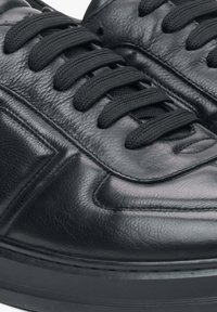 Černé kožené tenisky s texturovaným povrchem, vybavené černými plochými tkaničkami, šitými detaily a gumovou podrážkou. Detailní pohled na designové prvky.