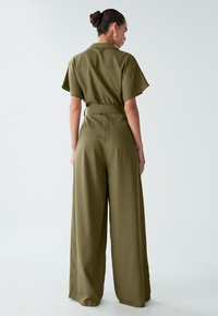 Oljčno zelena kombineza iz tkanine s gladko teksturo, z kratkimi rokavi, ovratnikom in širokimi hlačnicami z pasom na pasu.
