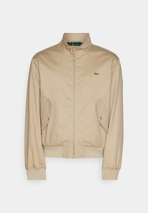 Chaqueta bomber beige con cuello alto, cierre de cremallera, bolsillos laterales en ángulo y puños de canalé. Presenta un logo verde en el pecho.