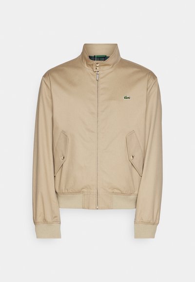 Veste bomber beige avec col montant, fermeture éclair, poches latérales obliques et poignets côtelés. Dispose d'un logo vert sur la poitrine.