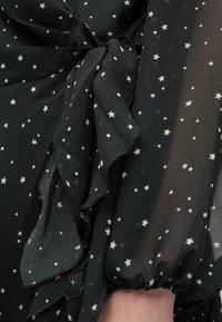 Sort gennemsigtigt stof med hvide stjerne-mønstre, bundet i en rynket sløjfe nær et håndled med en samlet mansjet. 