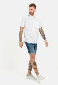 Hvid kortærmet skjorte med knapper og brystlomme, parret med denimshorts og hvide sneakers. Viser synlige tatoveringer på benene.