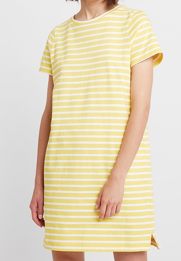 Nainen, joka pitää ylipitkistä, kyynärpäähän asti ulottuvista kelta-valkoisista vaakasuorista raidoista koostuvaa mekkoa, jossa on sivuhalkio, seisoo yksinkertaista taustaa vasten.