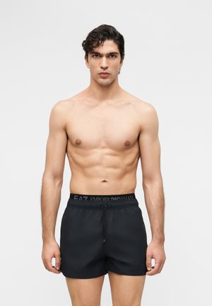 Skjorteløs mand med mørkt krøllet hår iført sorte EA7 Emporio Armani badeshorts, stående mod en ensfarvet lys grå baggrund.