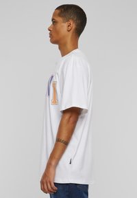 White cotton t-shirt featuring short sleeves and a crew neck. Front displays colorful "NY" text. Notable side slit detail at the hem.