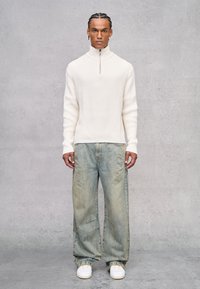 Maglione bianco a coste con zip, jeans ampi blu chiaro e sneakers bianche. Modello in piedi davanti a uno sfondo neutro in cemento.