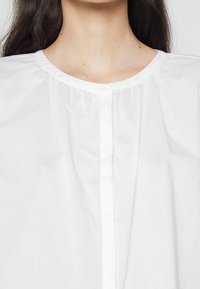 Chemise blanche à boutons avec un col rond, des détails froncés et une texture lisse. Comprend une couture centrale et une fermeture à un seul bouton.