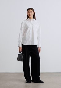 Λευκό βαμβακερό πουκάμισο με κουμπιά, με τσέπη. Συνδυασμένο με άνετο μαύρο παντελόνι και μικρό μαύρο τσαντάκι, πάνω σε ουδέτερο φόντο.