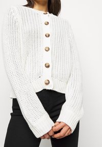 Cardigan blanc en tricot avec une coupe courte, de grands boutons ronds, une texture côtelée et de longues manches avec des larges poignets.
