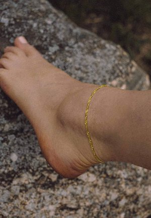Gold ankle bracelet on a bare foot, featuring a delicate chain design, against a textured stone background.