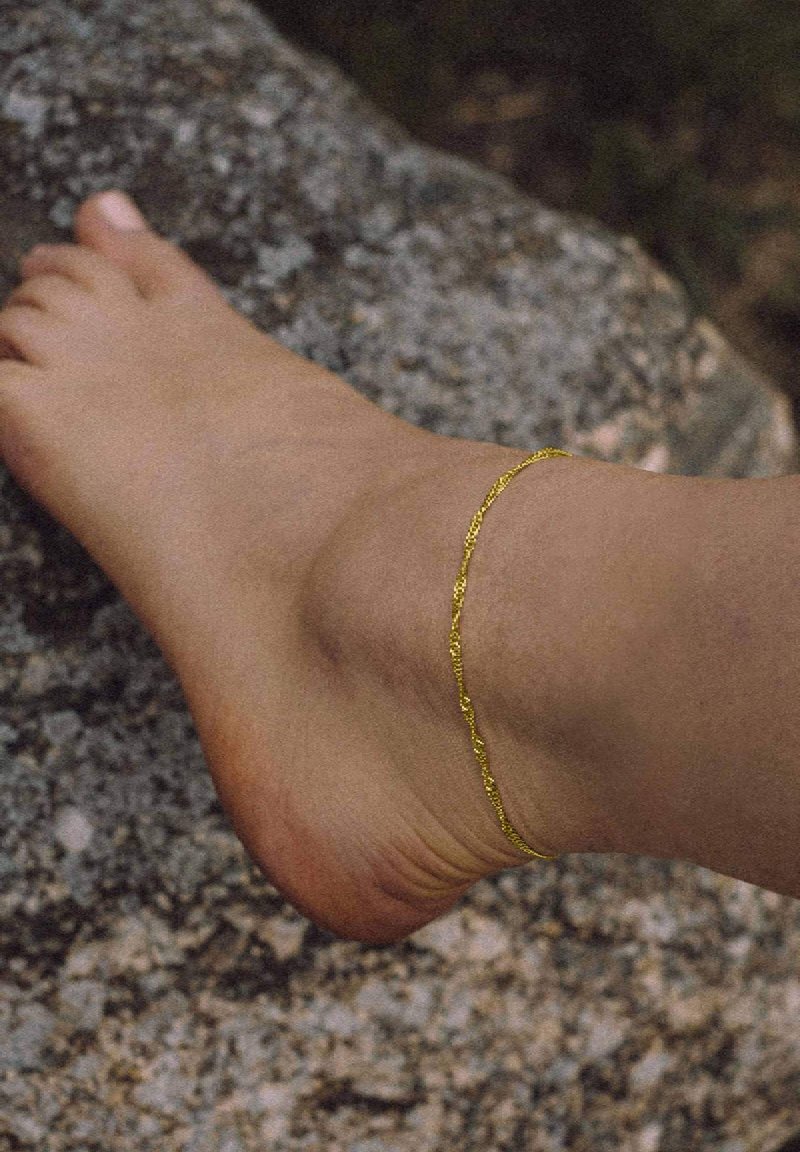 Gold ankle bracelet on a bare foot, featuring a delicate chain design, against a textured stone background.