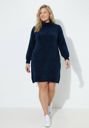 Vrouw loopt en draagt een marineblauwe gebreide jurk met lange mouwen en witte sneakers tegen een effen witte achtergrond.
