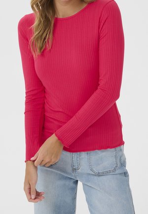 Vrouw draagt een aangesloten, geribbelde, lange mouwen rode top en lichtblauwe spijkerbroek, met één hand die de mouw aanpast.