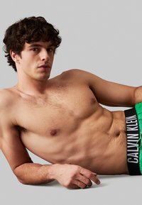 Boxer in cotone verde con un cinturino nero che presenta il testo bianco "CALVIN KLEIN". Il modello ha i capelli corti e ricci e un torso nudo con peli chiari.