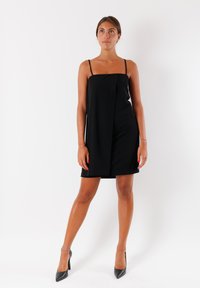 Vestito mini nero con spalline sottili, scollatura dritta, frontale drappeggiato e texture liscia. Abbinato a tacchi alti neri.
