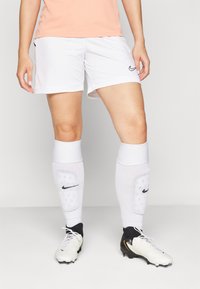 Nike spordilühikesed püksid valges, millel on elastne vöökoht ja must logo, koos valgete põlvikutega ja musta-valgete jalgpallijalatsitega.