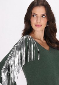 Pulover verde cu un design în formă de V, având franjuri din paiete argintii pe o umăr, evidențiind un aspect texturat și vibrant.