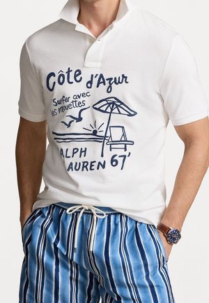 Biała koszulka polo z granatowym nadrukiem przedstawiającym scenę plażową i tekst. Krótkie rękawy, zapięcie na guziki, połączona z niebieskimi spodniami w paski.