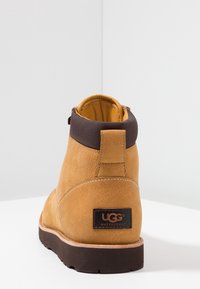 Botín de cuero UGG impermeable en color beige con cuello marrón oscuro y suela negra gruesa, visto desde la parte trasera sobre una superficie blanca.
