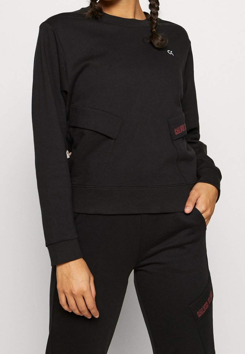 Femme en sweat-shirt noir à manches longues et pantalon assorti avec le logo rouge Calvin Klein sur les écussons de poche, debout avec une main dans la poche.