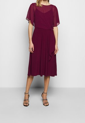 Kobieta ubrana w bordową sukienkę do kolan z przezroczystymi krótkimi rękawami i wiązaniem z boku, do której dopasowano czarne sandały na wysokim obcasie z paskami.