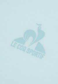 Světle modrá látka má jemně tištěné logo "Le Coq Sportif", které vyniká hladkou texturou a minimalistickým designem.