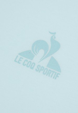 Světle modrá látka má jemně tištěné logo "Le Coq Sportif", které vyniká hladkou texturou a minimalistickým designem.