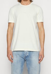 Krótki rękaw, kremowa koszulka wykonana z bawełny. Posiada okrągły dekolt i subtelne logo "BOSS" na środku. Gładka faktura.