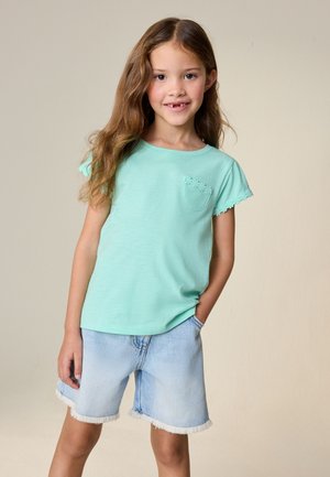 Ragazza giovane con lunghi capelli castani che indossa una camicia verde menta con tasca bordata da motivi floreali e pantaloncini di jeans chiari, in piedi con una mano in tasca.