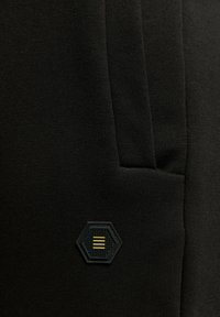 Zwarte joggingbroeken van stof hebben een getextureerd paneel en een hexagonale zwarte patch met drie gele strepen. Het ontwerp benadrukt een eenvoudige, slanke vorm.