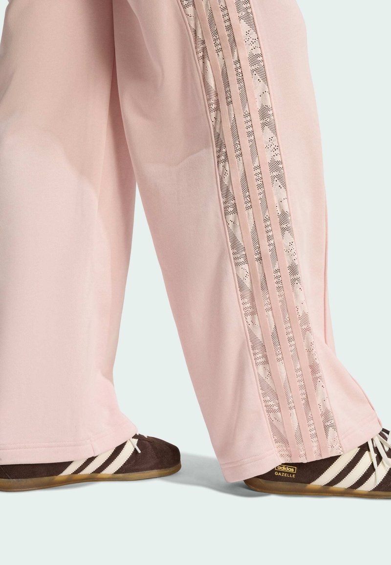 Light pink sweatpants feature a side panel with stripes and a decorative lace pattern. Dark brown sneakers with white stripes are worn.