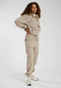 Beige sweatshirt med høj hals, parret med matchende sweatpants med elastisk talje. Modellen bærer hvide sneakers med detaljer.