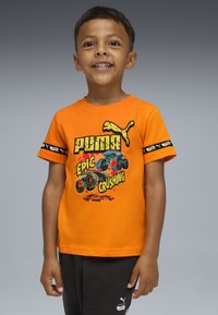 Ein lächelnder Junge trägt ein oranges Puma-T-Shirt mit Monster-Trucks und schwarze Hosen vor einem grauen Hintergrund.