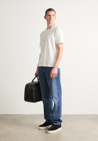 Witte katoenen t-shirt, blauwe spijkerbroek, zwarte leren tas en zwarte sneakers met een dikke witte zool, getoond op een staande mannelijke figuur.