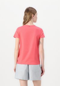 Korge t-skjorte med korte ermer i korallfarge, rund hals og en liten svart detalj på baksiden, brukt sammen med lyse grå shorts med lommedetaljer.