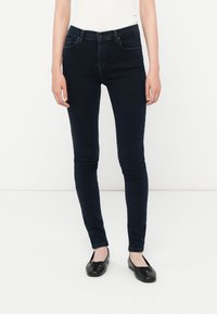 Sötétkék, skinny farmernadrág magas derékkal, rugalmas denim anyagból készült. Öt zsebből áll, sima textúrájú. Fekete balerina cipővel párosítva.