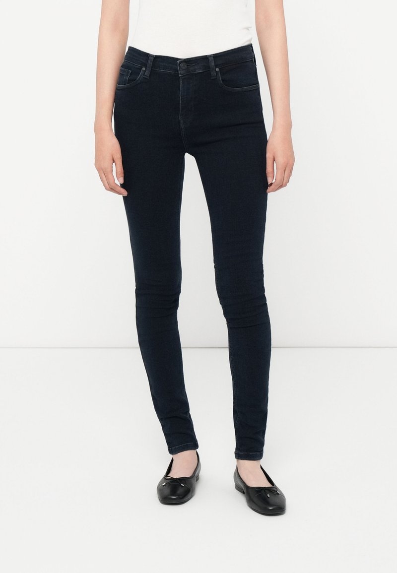 Sötétkék, skinny farmernadrág magas derékkal, rugalmas denim anyagból készült. Öt zsebből áll, sima textúrájú. Fekete balerina cipővel párosítva.