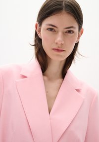 Gaiši rozā bleizeris ar izgriezumu un strukturētām plecu daļām. Audums šķiet gluds ar vieglu spīguļojumu. Priekšā ir V-veida izgriezums.