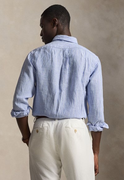 Άνδρας με την πλάτη στραμμένη φορώντας μπλε και λευκή ριγέ μακρυμάνικη πουκάμισο με γυρισμένα μανίκια και λευκό παντελόνι, με ουδέτερο φόντο.