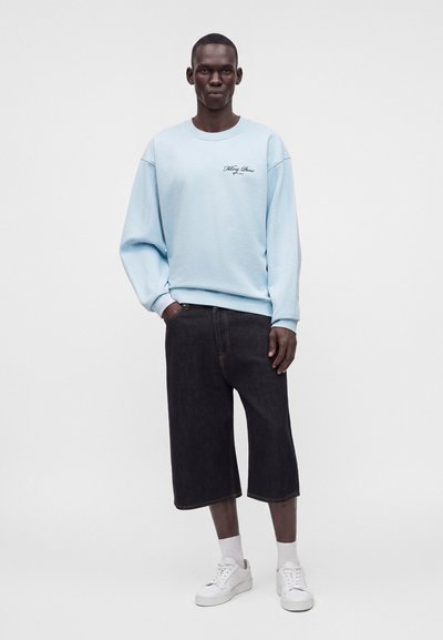 Homme portant un sweat-shirt bleu clair avec un petit texte, un short large noir jusqu'au genou, des chaussettes blanches et des baskets blanches, debout devant un fond uni.