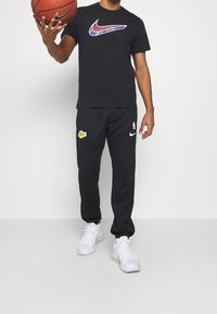 Camiseta negra de Nike con un logo multicolor, combinada con pantalones de chándal negros de Nike que tienen un logo de los Lakers, y zapatillas atléticas blancas.
