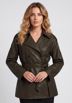 Frau mit welligem blondem Haar, die eine dunkelgrüne, taillierte Lederjacke trägt und vor einem schlichten hellgrauen Hintergrund steht.
