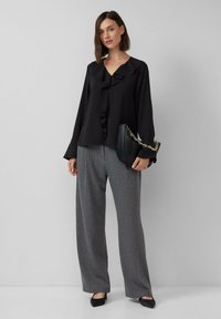 Zwarte blouse met lange mouwen en ruches op de kraag, gecombineerd met grijze gestreepte wijde broek en zwarte platte schoenen. Zwarte handtas met gouden ketting.