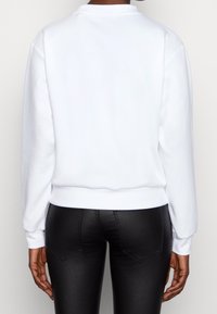 Felpa bianca con maniche lunghe, scollatura a girocollo, polsini e orlo a costine. Tessuto liscio, stile aderente, indossata con pantaloni neri attillati.
