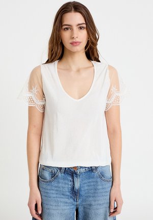 Junge Frau trägt ein weißes V-Ausschnitt-Top mit transparenten Spitzenshorts und blaue Jeans, steht vor einem schlichten hellen Hintergrund.