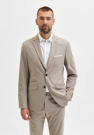 Traje gris claro con chaqueta de un solo botón, solapas de muesca y dos bolsillos frontales. Combinado con una camisa blanca y botones marrones.