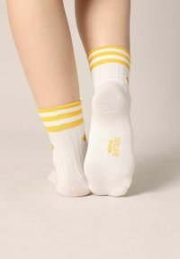 Witte sokken met gele strepen bij de boord en gele accenten. Heeft een geribbelde textuur en de tekst "FEEL LIKE WOMEN" gedrukt op de zool.