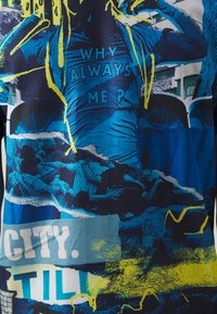 Grafische T-shirt met een blauw en zwart ontwerp met gele accenten, waarop de tekst "WHY ALWAYS ME ?" staat, evenals verschillende afbeeldingen en patronen.