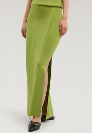 Mujer con una falda larga verde lima con una abertura frontal, combinada con zapatos planos negros de punta puntiaguda con detalle de hebilla dorada.